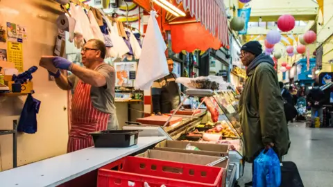 Getty Images A butcher in a red-and-white striped apron and blue gloves serves a customer at an indoor counter in Brixton. 