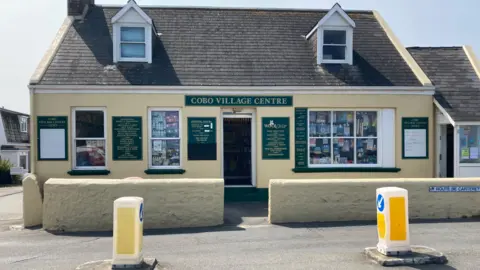 BBC A yellow house-like building with a green sign above the door that says "Cobo Village Centre". The photo is taken from the road. 