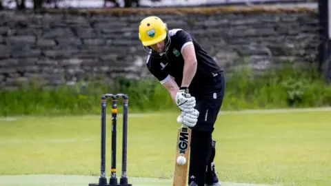 A man wears dark clothes, a yellow helmet and white gloves as he bats a ball in front of three cricket stumps.