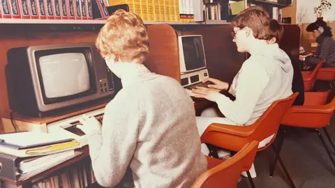An old colour picture of students in orange plastic chairs sitting at computer terminals from years gone by