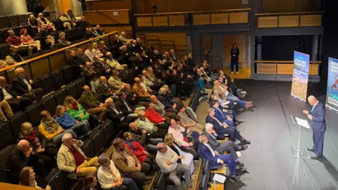BBC A man on a stage at a lectern giving a speech to an auditorium of people in raked seating. 