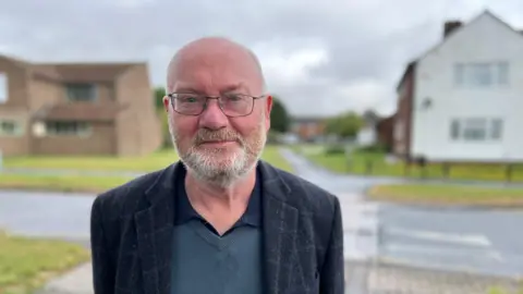 A bearded man stands in front of a road crossing. He is wearing a dark blue suit jacket with a lighter blue jumper and navy shirt underneath. He has rectangular, metal-framed glasses on. A few houses and a footpath can just be made out in the background, which has a blur effect on it.