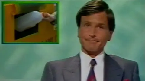 Peter Levy presenting in his earlier years. He has short brown hair and is wearing a grey suit jacket, a white shirt and blue and red tie. He is sitting in front of a green screen with an inset picture of a milk carton being deposited into a yellow bin.