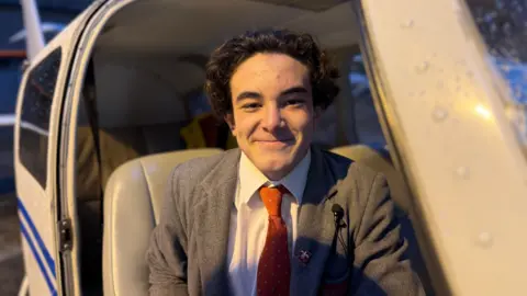 A young boy in the seat of a small plane. He is smiling at the camera. He is wearing a grey school blazer and a red tie.