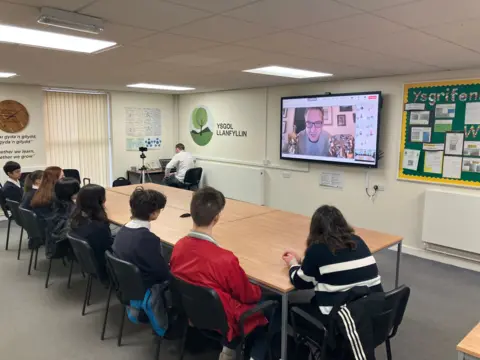 Pupils sat in classroom at a table. On the projector is a man speaking with him. In the frame of him you can see an Oscar and a BAFTA. The students are in uniform, most are in blue jumpers and white shirts. One man in the right of the image is wearing a red jacket, a woman next to him has a blue ad white striped jumper on. 