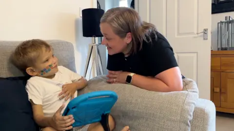 Harrison is playing on a blue tablet looking up at his mum Kirsty who is leaning on a sofa armrest.