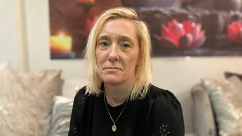 A woman with shoulder-length blonde hair, wearing a black top with gold necklace, sits on a sofa.