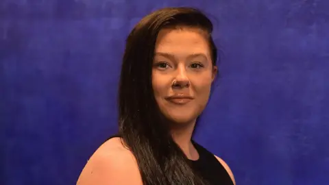 NI Equality Commission Ms Storey is standing in front of a blue backdrop. She has long brown hair and is wearing a black top and has a hoop piercing on her nose. 