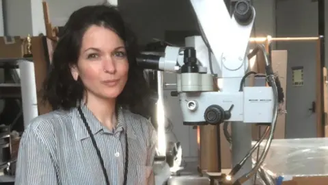 Northumbria University PhD student Jennifer Myers in a lab with a large microscope-type machine next to her. She has long black hair and is wearing a black and white striped blouse. She is slightly smiling at the camera.