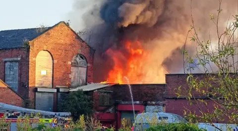 Fire tears through a red-brick building as smoke plumes. A fire engine is in the foreground and crews can be seen spraying water.