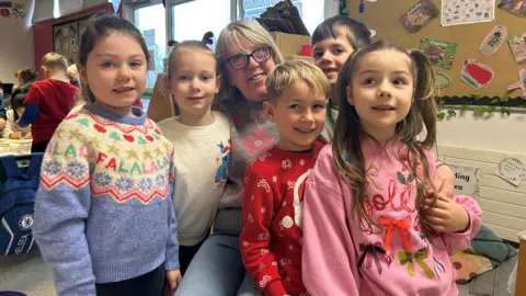 Gwen Rose smiling with five children. They are all wearing Christmas jumpers.