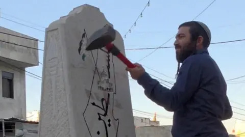 A man in a blue shirt hits a stone memorial with a sledgehammer