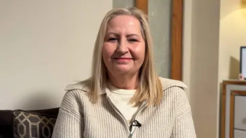 Kath Nixon is sitting in the living room of her home looking down the lens of the camera. She is smiling and has shoulder length blonde hair. She is wearing an oatmeal fine knitted zipped top with large collar.
