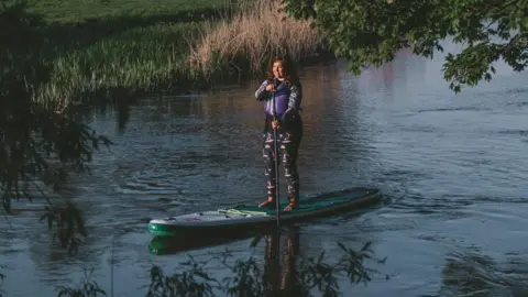 VIMA Photography A woman with dark hair stands on a paddleboard in a purple life jacket. It appears to be dusk and the water is dark and still.