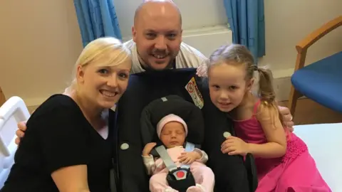 Handout Kaley and her husband David smile at the camera as their baby sits in a car seat and a blonde girl in a pink dress sits next to her.