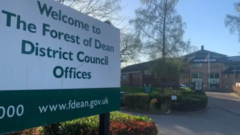 A sign which says 'Welcome to The Forest of Dean District Council Offices' in green writing on a white background, outside a large council building. There is a flagpole with a Union Jack on it, and a number of hedges and trees. 