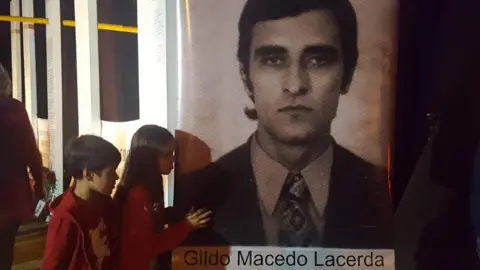 Tessa Moura Lacerda/Family handout Two children standing to the left of a black and white picture of a man