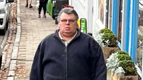 BBC A man with greyish black hair slicked back and glasses in a blue zip up top and beige trousers walks along a cobbled street with shops to one side