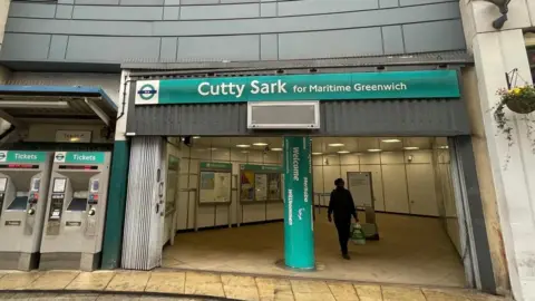 Front entrance of Cutty Sark station in Greenwich. The picture shows two ticket mchines and the entrance hall during daylight, with one person walking in