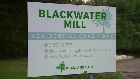 BBC A pale green sign that says 'Blackwater Mill' in large green writing. Behind it is a car park and a grass lawn.