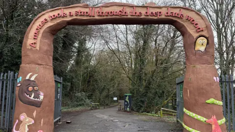 A tall brown archway marking the entrance to the Gruffalo Trail in Colin Glen Forest Park. It is colourfully decorated with animals including a mouse, a fox, an owl, a snake and the Gruffalo. A sign across the top of the arch reads: "A mouse took a stroll through the deep dark wood".