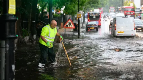 Getty Images A council worker in a high viz jackets tries to tackle a flooded road in London. He is using his broom to try to clear a drain. Cars and buses are driving past through the flood waters.