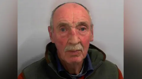 A man with balding hair and moustache looking directly ahead. He is wearing a green hooded top with a blue shirt underneath.