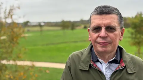 Mike Jelfs, wearing a wax jacket with a shirt underneath stands on a bridge in Whetstone. In the background a dog-walking park can be seen - further into the distance farmland earmarked for a large housing development known as Whetstone Pastures.