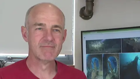 Dom Robinson - wearing a red T-shirt and has grey hair and a bald head. A computer screen with diving footage is on the screen behind him