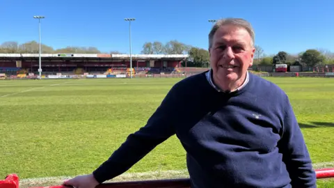 Steve Durham. He has short white hair and is wearing a blue jumper. He is standing in front of the grass pitch, leaning against a red metal railing. A low stand is on the other side of the Borough Park pitch.