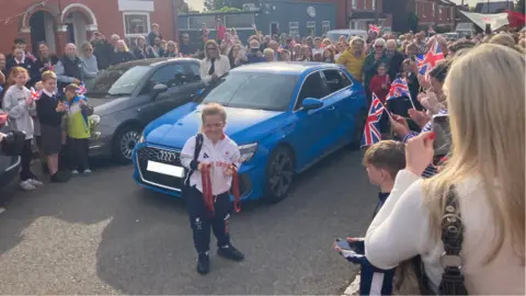 James Burridge/BBC Maise Summers-Newton standing in front of a blue car while holding three medals, two gold and one bronze, and smiling.
She is surrounded by people waving Union Jack flags and smiling to greet her as she comes home.