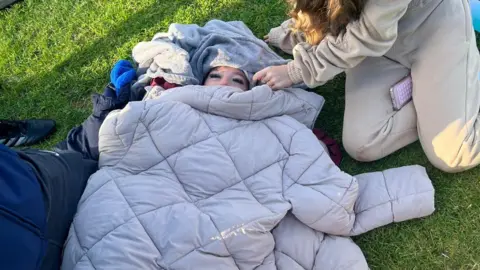 Charlene Paterson Jackets cover a woman lying on the ground and only her eyes can be seen. She is lying on the ground with two people kneeling around her
