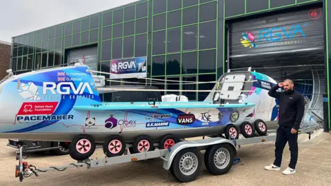 Oliver Rathbone/George Attfield George, dressed in black with white trainers, stands next to their boat, Pacemaker, which is on a trailer