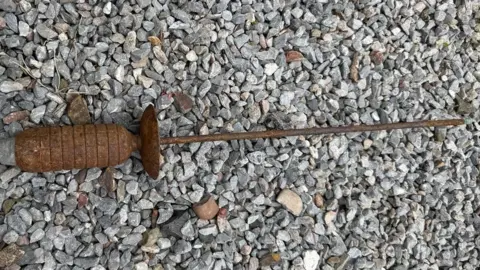 A rusted, cylindrical metal object with ridges and a long thin rod attached lies on a bed of small grey stones. The object appears weathered and corroded. The toe of a dark shoe is visible in the top-left corner, providing a sense of scale.
