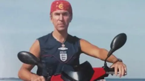 Mris Ile wears a red bandana on his head. He is astride a red and black motorbike and is wearing an England football team navy vest. 