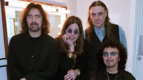 Butler, Osbourne, Ward and Iommi, dressed in black, sitting around a lit dressing room mirror. Their expressions are solemn as they look at the camera.