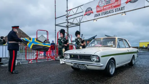 A man wearing a uniform waves the Caithness flag to signal a classic Ford car's departure from John O' Groats. Two pipers stand playing a tune at the start line.