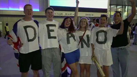 A group of Tom Dean's friends and family. They have their arms around each other and are smiling. They are wearing white t-shirts which spell out 'Deano'