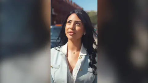 A woman with long black hair, in a light blue suit, looks up while walking in the street