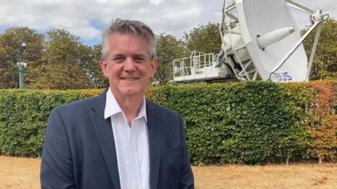 A man is wearing a smart blue suit and white shirt. He is standing next to a satellite, smiling.