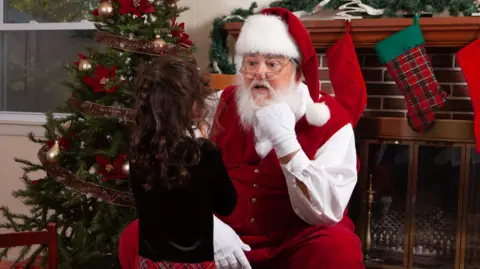 Father Christmas is wearing a red hat with white fur trim and white pom pom. He has a white shirt, white gloves and a red waistcoat and trousers. He is wearing glasses. Behind him is a mantelpiece with red and green stocking hung up, and there is a Christmas tree next to it. In front of him, with her back to the camera is a young girl with curly brown hair and a black top.