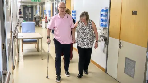 Norman and Christine taken in a hospital corridor. Norman is walking with a walking stick helped by Christine at his left side. He is bald-headed and is wearing glasses with a yellow tint. He has a pink t-shirt. Christine has short blonde hair and is wearing a sleeveless patterned shirt. She is displaying a watch on her arm. A table and chair in to the side of the corridor behind them and there is various medical equipment along the walls.