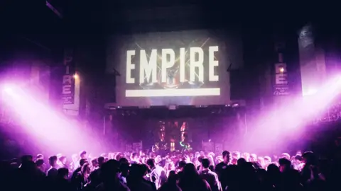 Inside of a nightclub. The dance floor is packed full with people, and purple strobe lights shine down on them from the right and left. Above the dance floor is a screen that says 'EMPIRE' in white.