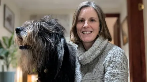 Kirstie Pickles is on the right next to a large, shaggy dog with its head turned away from her. She has shoulder-length grey hair, is smiling and looking at the camera, wearing a grey knitted top.