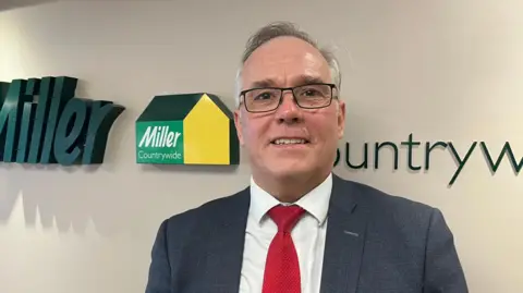 A man wearing a blue suit, white short and red tie, wearing glasses