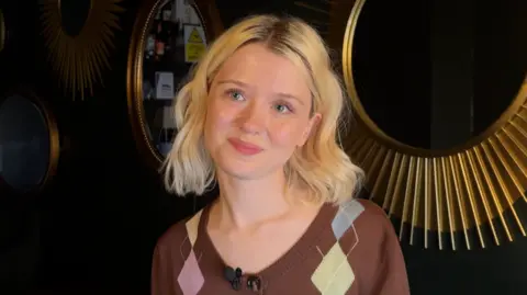 A young women with shoulder length blonde hair is smiling. She is wearing a brown cardigan and standing against a wall with two gold mirrors on it