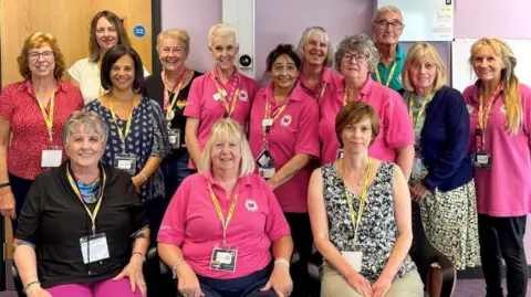 Great Western Hospital A group of volunteers smile for a picture. Many of them are wearing a pink branded polo shirt, and one male volunteer weas the same shirt in a teal colour. All of them wear a yellow lanyard, and appear to be posing inside a hospital. 