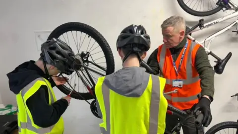 Thomas and Travis wearing hi-vis jackets working on a bike that is on a stand. Danny Bryce is also working on the bike and is wearing an orange hi-vis jacket.