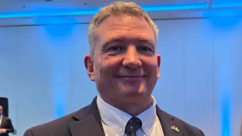 Derek Roberts is wearing a dark suit and a white shirt.