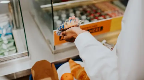 Getty Images A stock image of a person holding a wrapped item of food in their hands while at a canteen station. In the background are blurred stations with drinks and snacks.